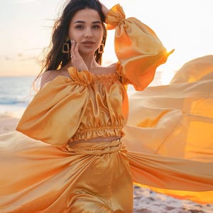May include: A golden-yellow off-the-shoulder dress with long, billowing sleeves. The dress has a cinched waist and a flowing skirt, creating a dramatic silhouette. The model is wearing gold earrings. The background is a beach at sunset.