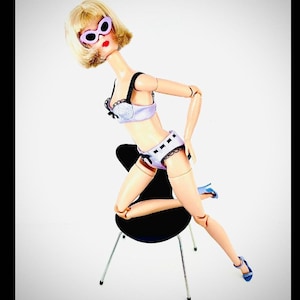 May include: A doll wearing a lavender lingerie set with black trim, matching sunglasses, and blue heels. The doll is posed on a black chair with silver legs, set against a white background.