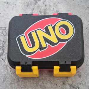 May include: A rectangular black plastic storage case featuring the word UNO in bold yellow letters set against a red oval background. The case has a textured surface and two bright yellow locking latches on the front edge for secure closure.