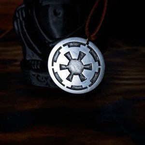 Pode incluir: Um colar com um pingente de prata com o símbolo imperial preto e branco de Star Wars.
