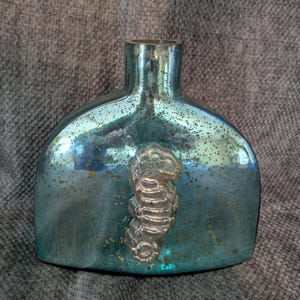 May include: A blue glass vase with a silver seahorse design. The vase has a textured surface and a rounded shape.