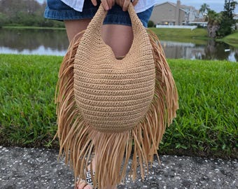 May include: A tan crocheted tote bag with fringe trim. The bag is made of natural fibers and has a round shape.