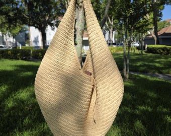 May include: A large, beige woven tote bag with a single shoulder strap. The bag is made of a textured, natural-looking material and has a rounded shape. A small, brown leather tag is attached to the side. The bag is hanging from a tree branch.