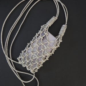 May include: A silver-toned, knotted cord phone pouch featuring a lattice-style macrame design. The pouch includes a long, integrated crossbody strap and holds a light-colored smartphone with a visible circular magnetic charging ring on the back.