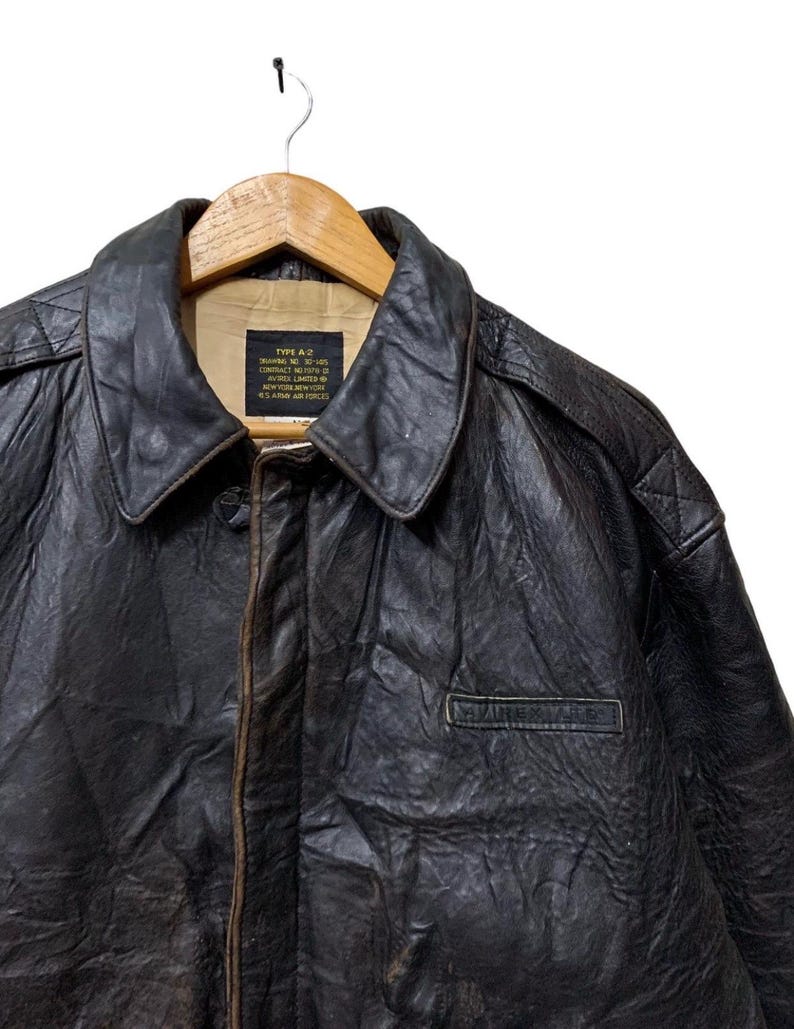 May include: A vintage brown leather Type A-2 bomber jacket with a collared neckline and a small pocket. The jacket is hanging on a wooden hanger. The text on the label reads "TYPE A-2" and "U.S. AIR FORCES".