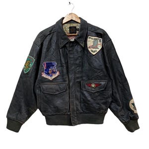 Może przedstawiać: Vintage czarna sk&oacute;rzana kurtka bomberka z wieloma naszywkami. Kurtka ma kołnierz, dwie przednie kieszenie i prążkowane mankiety. Naszywki obejmują emblematy i tekst w stylu wojskowym. Kurtka wisi na drewnianym wieszaku.