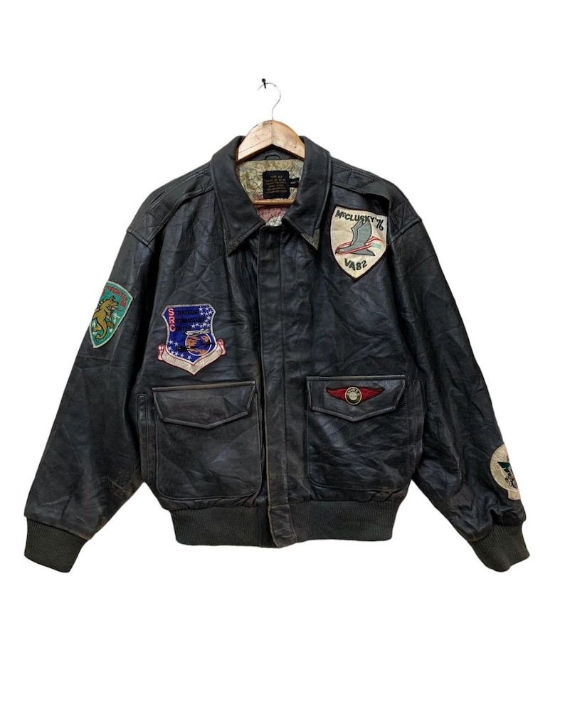 Może przedstawiać: Vintage czarna sk&oacute;rzana kurtka bomberka z wieloma naszywkami. Kurtka ma kołnierz, dwie przednie kieszenie i prążkowane mankiety. Naszywki obejmują emblematy i tekst w stylu wojskowym. Kurtka wisi na drewnianym wieszaku.