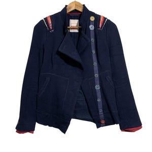 May include: A navy blue jacket with a unique design, featuring decorative buttons and colourful accents on the shoulders. The jacket has a tailored fit and a distinctive asymmetrical closure. The cuffs are lined with red fabric.
