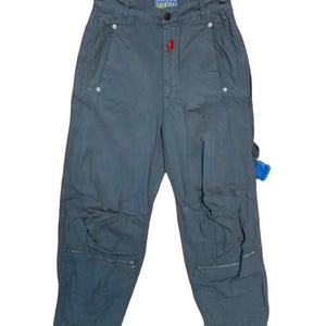 May include: Dark gray pants with multiple pockets and zippers. The pants have a high waist and a red accent near the button. The word "World" is visible on the waistband. The pants are hanging on a wooden hanger.
