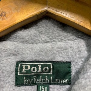 Pu&ograve; includere: Primo piano di un tessuto in pile grigio con un'etichetta verde. L'etichetta recita "Polo by Ralph Lauren 150" in lettere bianche. Il tessuto &egrave; drappeggiato su una gruccia di legno.