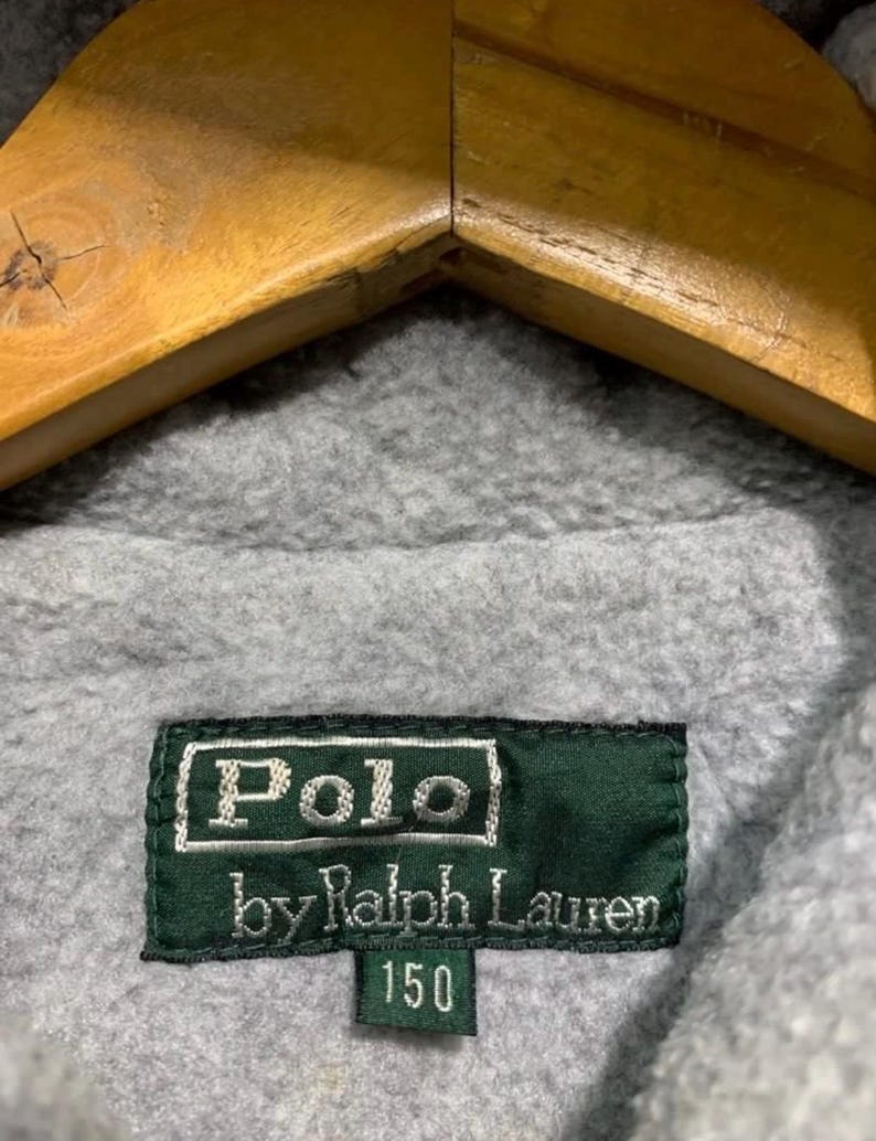 Pu&ograve; includere: Primo piano di un tessuto in pile grigio con un'etichetta verde. L'etichetta recita "Polo by Ralph Lauren 150" in lettere bianche. Il tessuto &egrave; drappeggiato su una gruccia di legno.
