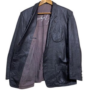 May include: Black leather blazer with a textured finish, hanging on a wooden hanger. The blazer has a classic design with a notched lapel, two front pockets, and a button closure. The interior lining is a muted purple color, with white text visible.