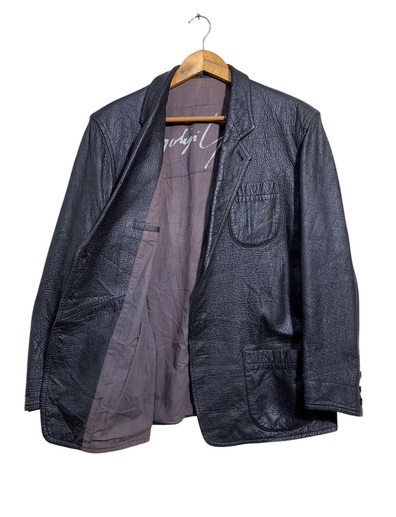 May include: Black leather blazer with a textured finish, hanging on a wooden hanger. The blazer has a classic design with a notched lapel, two front pockets, and a button closure. The interior lining is a muted purple color, with white text visible.