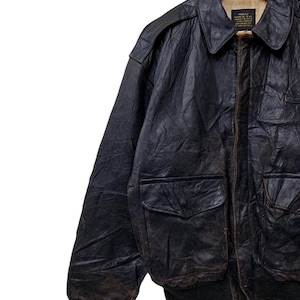 May include: A vintage black leather A-2 bomber jacket with two front pockets, a ribbed hem, and a buttoned collar. The jacket is hanging on a wooden hanger. The leather has a worn, distressed appearance.