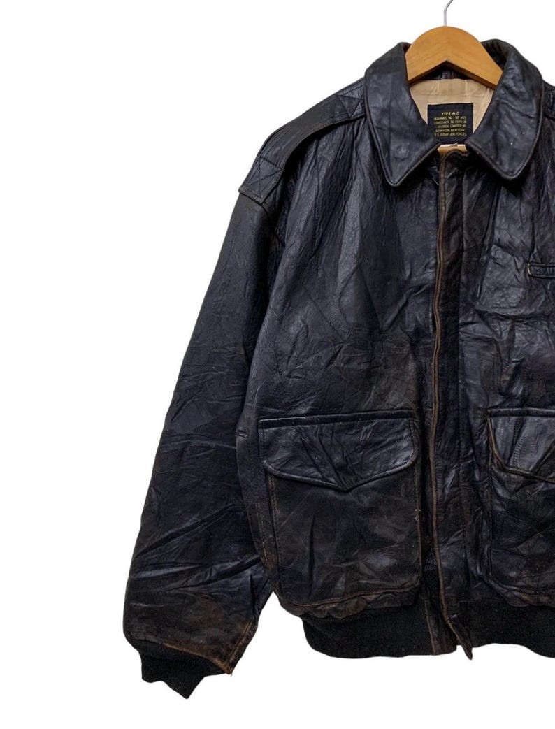 May include: A vintage black leather A-2 bomber jacket with two front pockets, a ribbed hem, and a buttoned collar. The jacket is hanging on a wooden hanger. The leather has a worn, distressed appearance.
