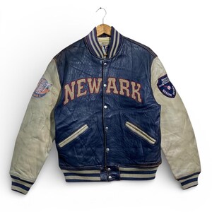 Puede incluir: Chaqueta universitaria vintage azul marino y crema. La chaqueta presenta la palabra "NEWARK" en letras rojas en el pecho. Las mangas son de color crema y tiene parches en las mangas. La chaqueta est&aacute; colgada en una percha de madera.