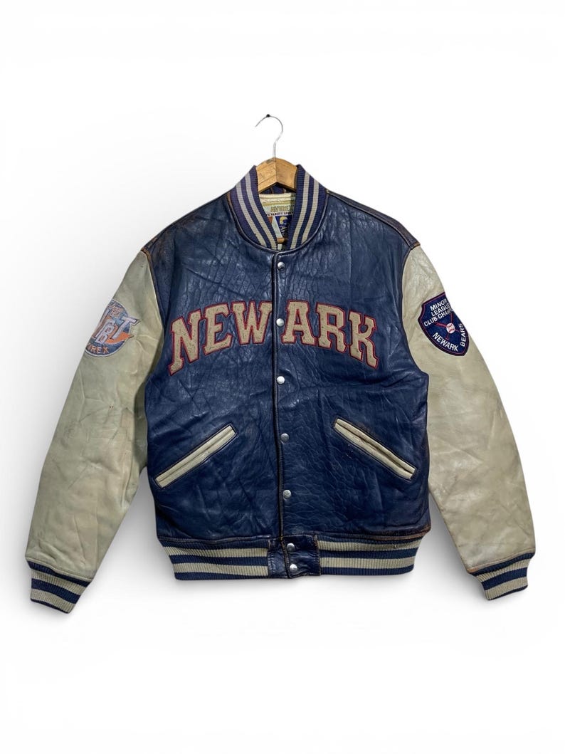 Puede incluir: Chaqueta universitaria vintage azul marino y crema. La chaqueta presenta la palabra "NEWARK" en letras rojas en el pecho. Las mangas son de color crema y tiene parches en las mangas. La chaqueta est&aacute; colgada en una percha de madera.