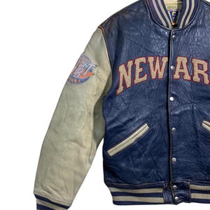 Puede incluir: Chaqueta universitaria vintage azul y crema con la palabra "NEW AR" en letras grandes de color canela en el pecho. La manga izquierda presenta un parche con texto. La chaqueta tiene cuello, pu&ntilde;os y dobladillo acanalados con rayas azules y crema.