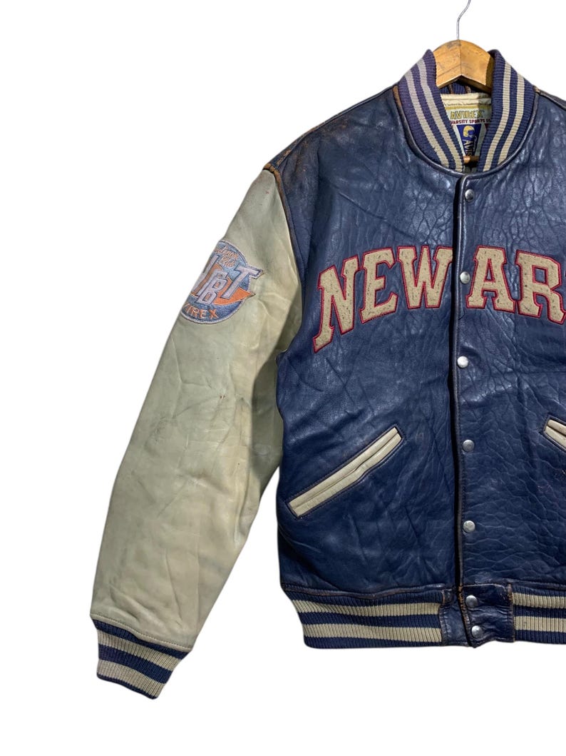 Puede incluir: Chaqueta universitaria vintage azul y crema con la palabra "NEW AR" en letras grandes de color canela en el pecho. La manga izquierda presenta un parche con texto. La chaqueta tiene cuello, pu&ntilde;os y dobladillo acanalados con rayas azules y crema.