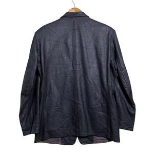 May include: A black leather blazer hangs on a wooden hanger against a white background. The blazer has a textured surface and a classic two-button design. The back view shows the tailored seams and the overall shape of the jacket.