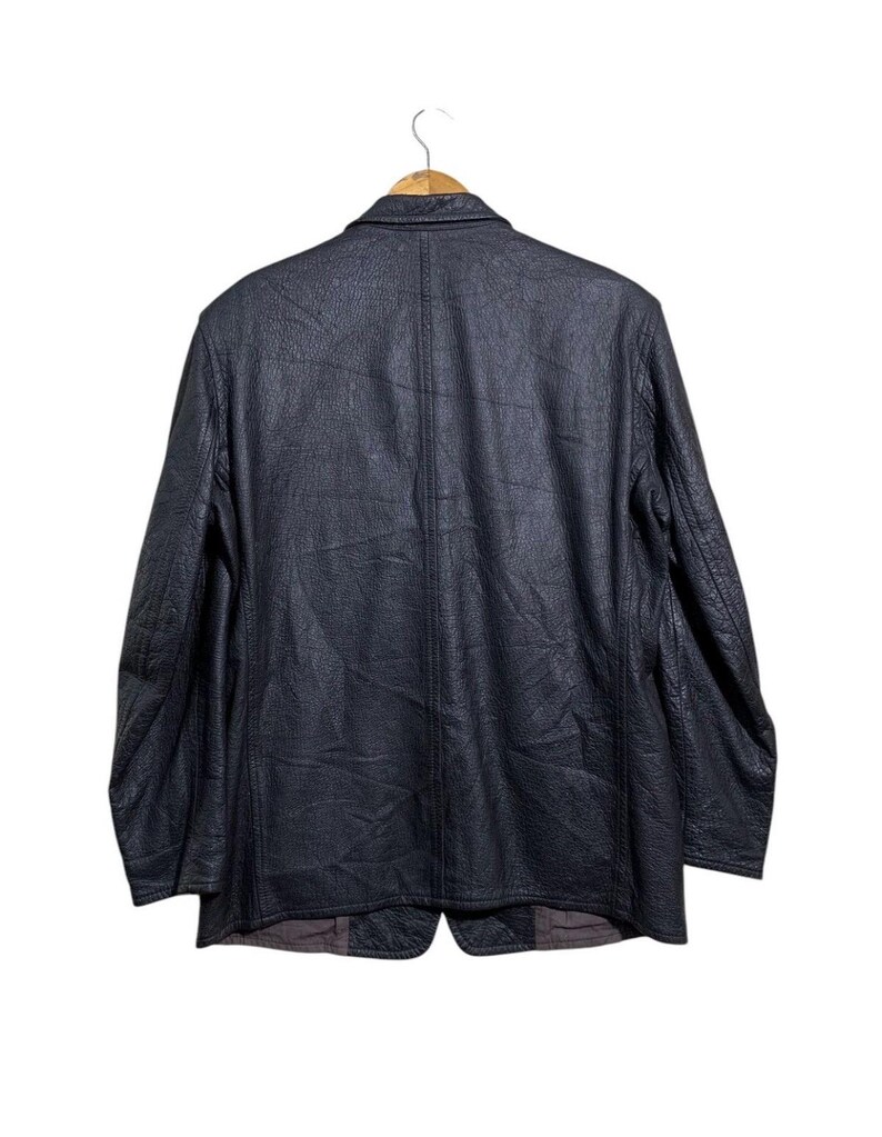 May include: A black leather blazer hangs on a wooden hanger against a white background. The blazer has a textured surface and a classic two-button design. The back view shows the tailored seams and the overall shape of the jacket.