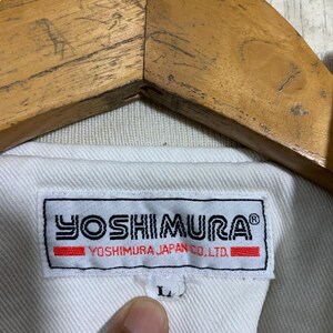 May include: Close-up of a white jacket with a label that reads "YOSHIMURA" in black and red. The label also includes "YOSHIMURA JAPAN CO., LTD." The jacket is hanging on a wooden hanger. The size is labeled as "L".