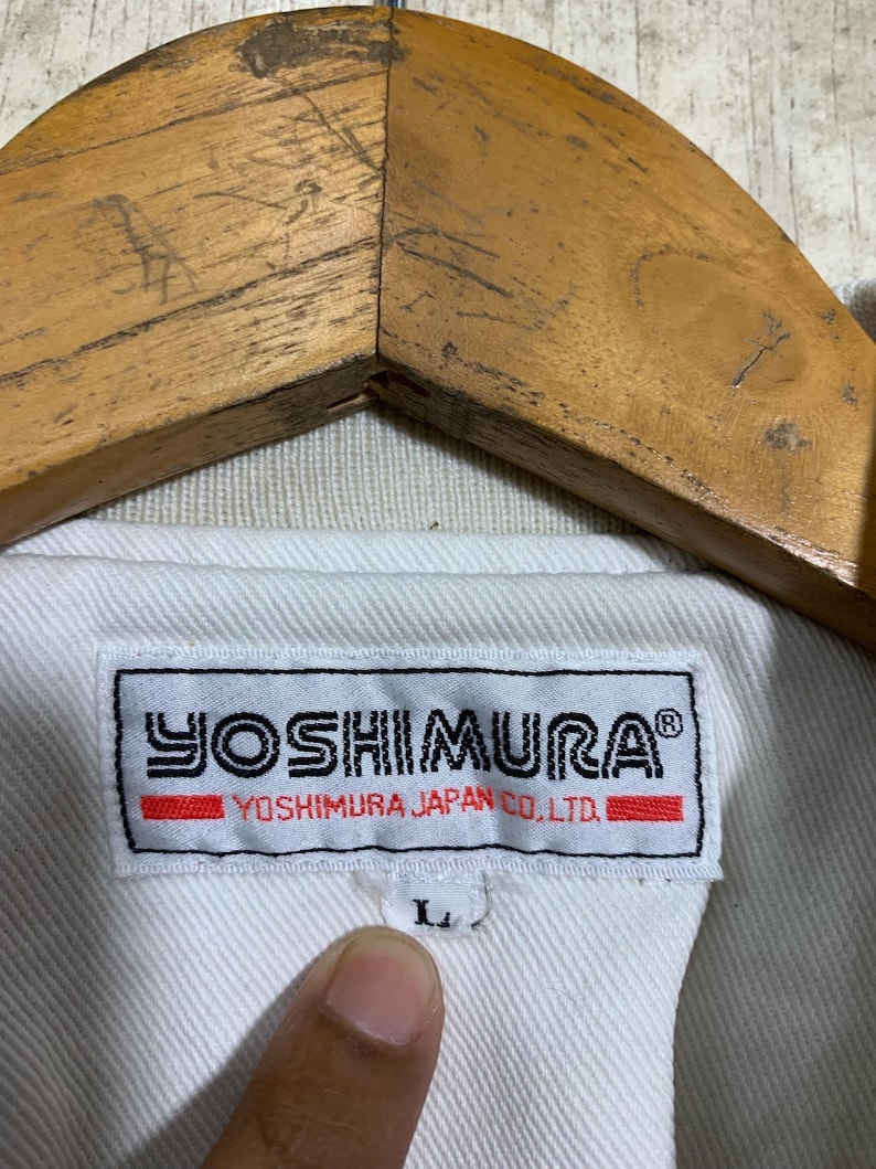 May include: Close-up of a white jacket with a label that reads "YOSHIMURA" in black and red. The label also includes "YOSHIMURA JAPAN CO., LTD." The jacket is hanging on a wooden hanger. The size is labeled as "L".