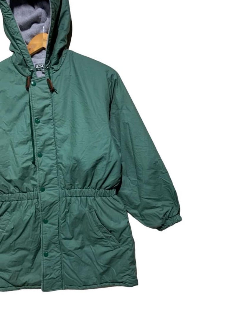 Pu&ograve; includere: Giacca con cappuccio verde con vita con coulisse e chiusura con bottoni. La giacca ha un cappuccio foderato in pile grigio e due tasche anteriori. La giacca &egrave; realizzata in un materiale leggero e sembra essere vintage.