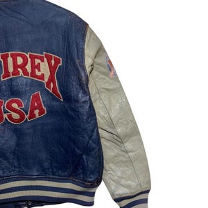 Puede incluir: Chaqueta de cuero azul y crema vintage con la palabra "AIREX USA" en letras rojas. La chaqueta tiene cuello y pu&ntilde;os a rayas, y un parche en la manga.