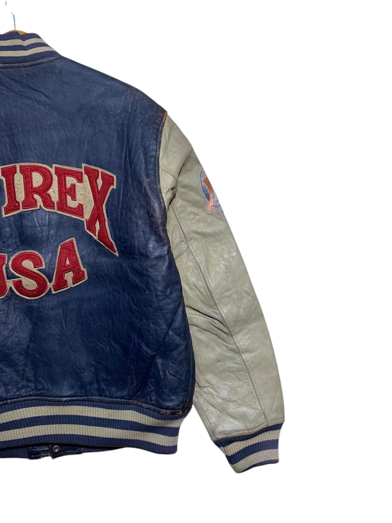 Puede incluir: Chaqueta de cuero azul y crema vintage con la palabra "AIREX USA" en letras rojas. La chaqueta tiene cuello y pu&ntilde;os a rayas, y un parche en la manga.