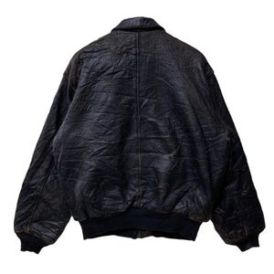 May include: A vintage black leather bomber jacket with a ribbed collar, cuffs, and hem. The jacket has a classic design with a slightly distressed appearance, suggesting it's been well-worn. The back view shows the full shape and texture of the leather.