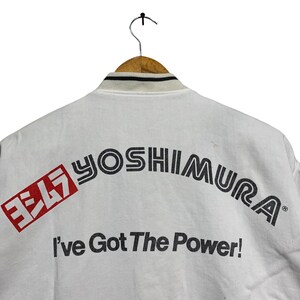 May include: White jacket with the text "YOSHIMURA" in black and red, and the phrase "I've Got The Power!". The jacket has a black and white striped collar and is hanging on a wooden hanger.