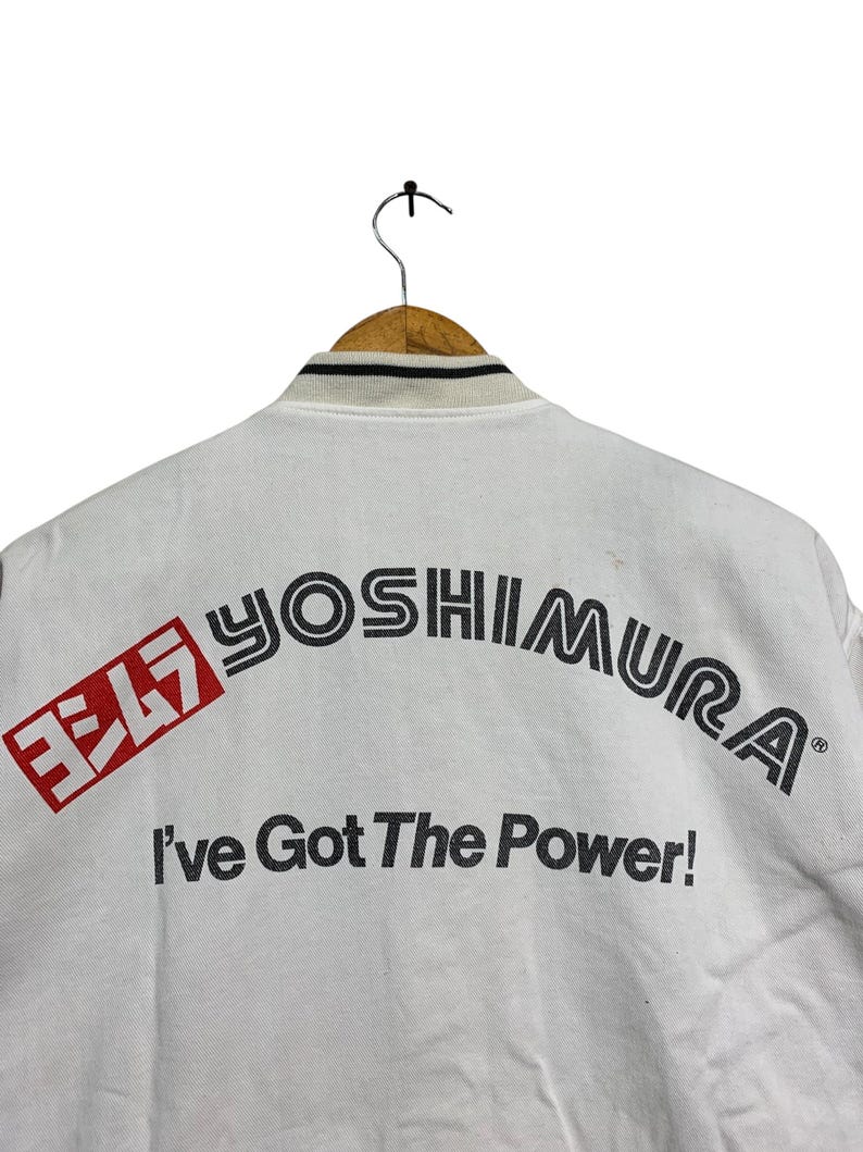 May include: White jacket with the text "YOSHIMURA" in black and red, and the phrase "I've Got The Power!". The jacket has a black and white striped collar and is hanging on a wooden hanger.