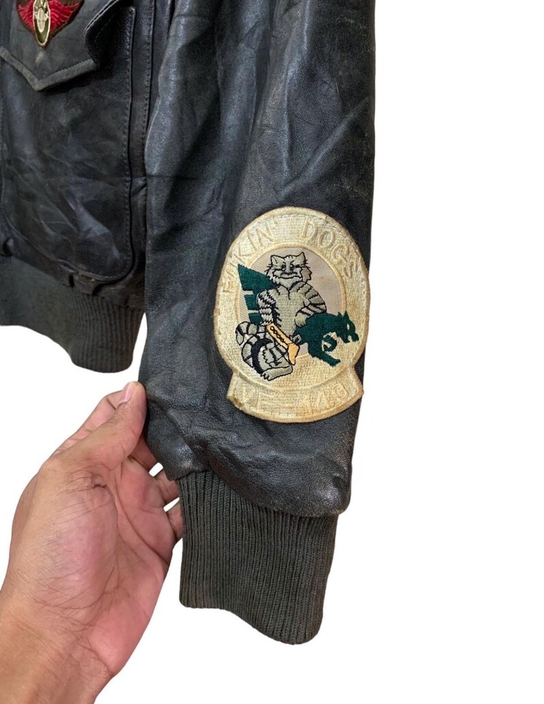 Może przedstawiać: Zbliżenie na czarną sk&oacute;rzaną kurtkę w stylu vintage z naszywkami. Kurtka posiada prążkowany mankiet i naszywkę z kresk&oacute;wkowym kotem i psem. Kurtka ma militarny styl.