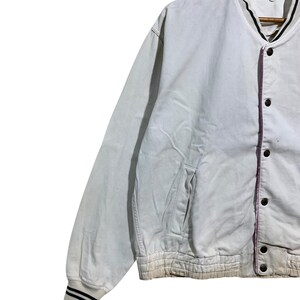 May include: A light-colored, vintage Yoshimura bomber jacket. The jacket features a button-up front, a ribbed hem and cuffs with black stripes, and a small pocket. The collar and trim are black and white. The jacket has the Yoshimura logo.