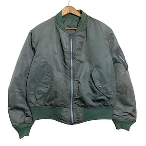 May include: Olive green bomber jacket with a green ribbed collar, cuffs, and waistband. The jacket features a silver zip, two front pockets, and a small pocket on the left sleeve. It hangs on a wooden hanger.