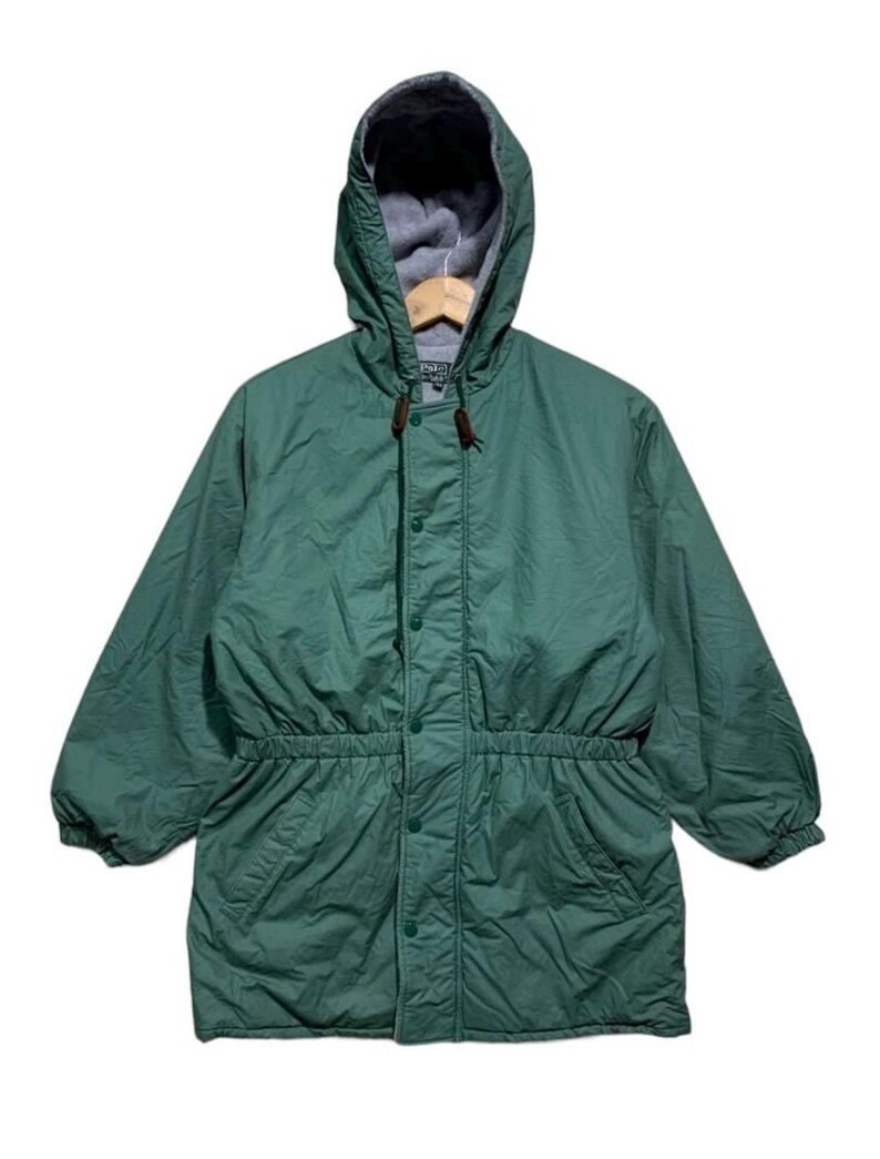 Pu&ograve; includere: Giacca verde con cappuccio, vita con coulisse e chiusura con bottoni. La giacca ha una fodera in pile grigio nel cappuccio ed &egrave; appesa a una gruccia di legno. La giacca ha due tasche anteriori.