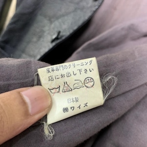 May include: Close-up of a garment tag with Japanese text and care symbols. The tag is white with black text and is attached to a gray fabric. The garment appears to be a jacket or coat.