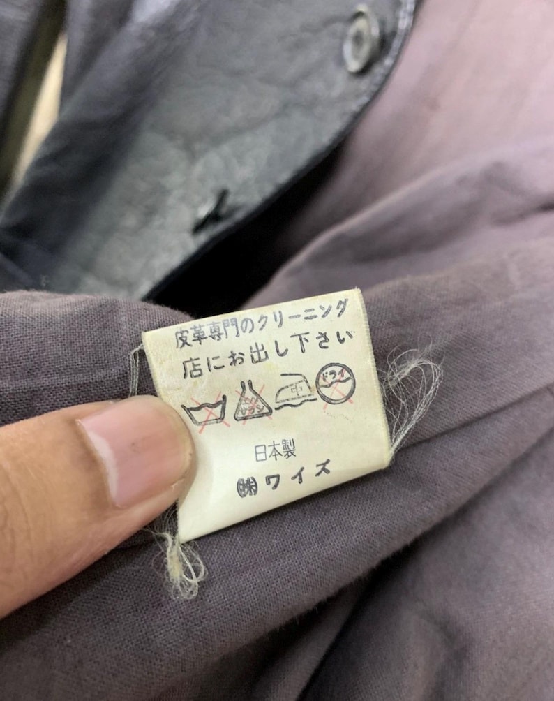 May include: Close-up of a garment tag with Japanese text and care symbols. The tag is white with black text and is attached to a gray fabric. The garment appears to be a jacket or coat.