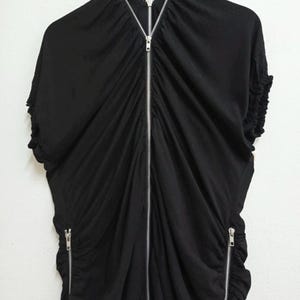 May include: Black dress with a draped design and a silver zipper running down the front. The dress has short sleeves and two additional zippers on the sides. The fabric appears to be soft and flowing, with gathered details.