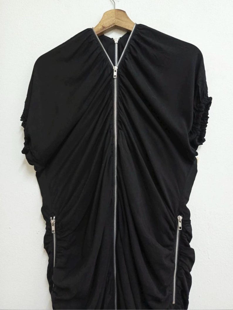 May include: Black dress with a draped design and a silver zipper running down the front. The dress has short sleeves and two additional zippers on the sides. The fabric appears to be soft and flowing, with gathered details.