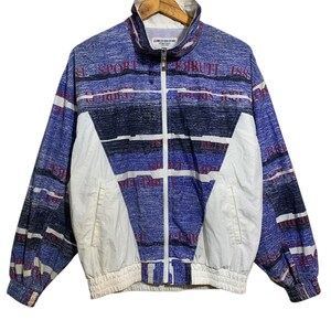 Könnte beinhalten: Eine Vintage-Sportjacke in Blau, Weiß und Rot mit durchgehendem Reißverschluss. Sie hat einen hohen Kragen und elastische Bündchen und Bund. Der Text "CIRUTE 188" ist auf Brust und Ärmeln aufgedruckt. Die Jacke hängt an einem Holzkleiderbügel.