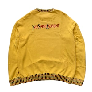 May include: Mustard yellow sweatshirt featuring the embroidered text "YVES SAINT LAURENT" in black and red. The collar and cuffs have a patterned trim of black, red, and yellow squares. The sweatshirt is a solid mustard yellow colour.