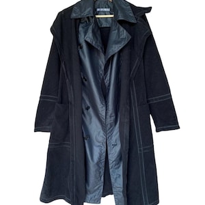 May include: Black long coat with a layered design. The coat features a double-breasted front, a notched collar, and a hood. The coat has a contrasting inner layer and visible stitching details. The coat is hanging on a wooden hanger.