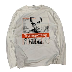 以下が含まれることがあります： 映画「Trainspotting」のグラフィックがプリントされた白い長袖Tシャツ。デザインにはポートレート、オレンジ色の映画タイトル、ミュージシャンリストが含まれています。シャツは柔らかく快適な素材で作られています。