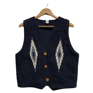 Op de afbeelding: Een donkerblauw mouwloos vest met een getextureerde gebreide stof en drie ronde houten knopen aan de voorzijde. Twee symmetrische ruitvormige patronen in tinten crème, lichtblauw en grijs zijn op het borstgedeelte geplaatst.