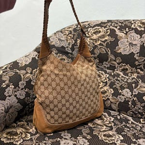 May include: A tan and brown shoulder bag with a repeating logo pattern. The bag has a brown leather strap and trim, with visible stitching details. The bag is sitting on a floral patterned surface.