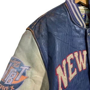 Puede incluir: Una chaqueta universitaria vintage azul y crema con la palabra "NEW" en letras grandes de color blanquecino con contorno rojo. La chaqueta presenta un parche con el texto "League Club" y "AVIREX". La chaqueta est&aacute; colgada en una percha de madera.