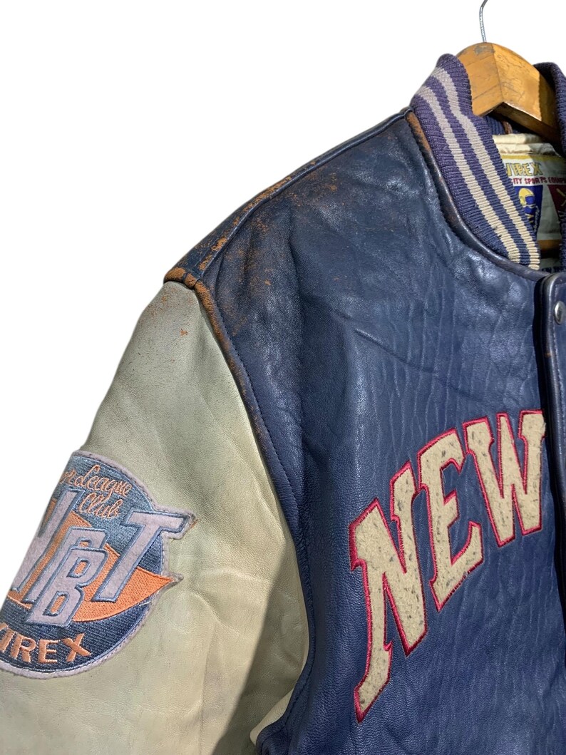Puede incluir: Una chaqueta universitaria vintage azul y crema con la palabra "NEW" en letras grandes de color blanquecino con contorno rojo. La chaqueta presenta un parche con el texto "League Club" y "AVIREX". La chaqueta est&aacute; colgada en una percha de madera.