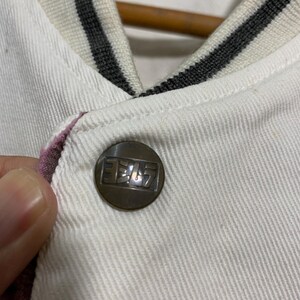May include: Close-up of a white jacket with a button. The button is bronze-colored with Japanese text. The jacket has a striped collar in white, gray, and black. The edge of the jacket is lined with a pink fabric.